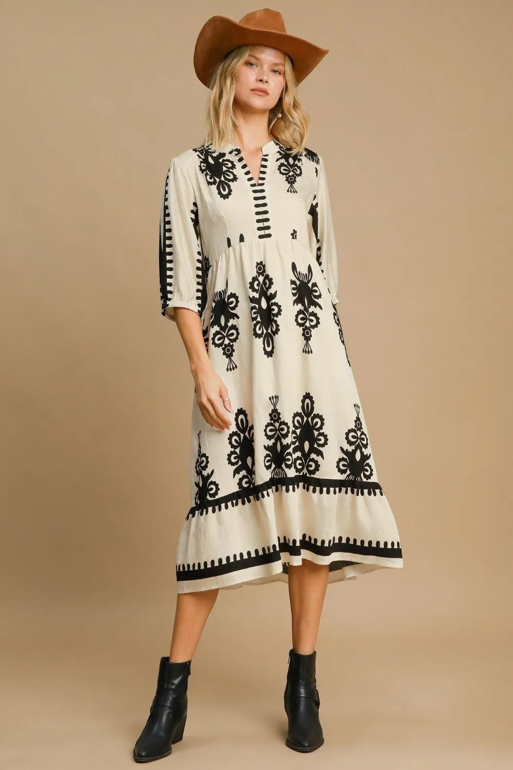 Stylio cream midi dress with black damask print, notched neckline, ruffled hem, and 3/4 sleeves. Model wears a cowboy hat.